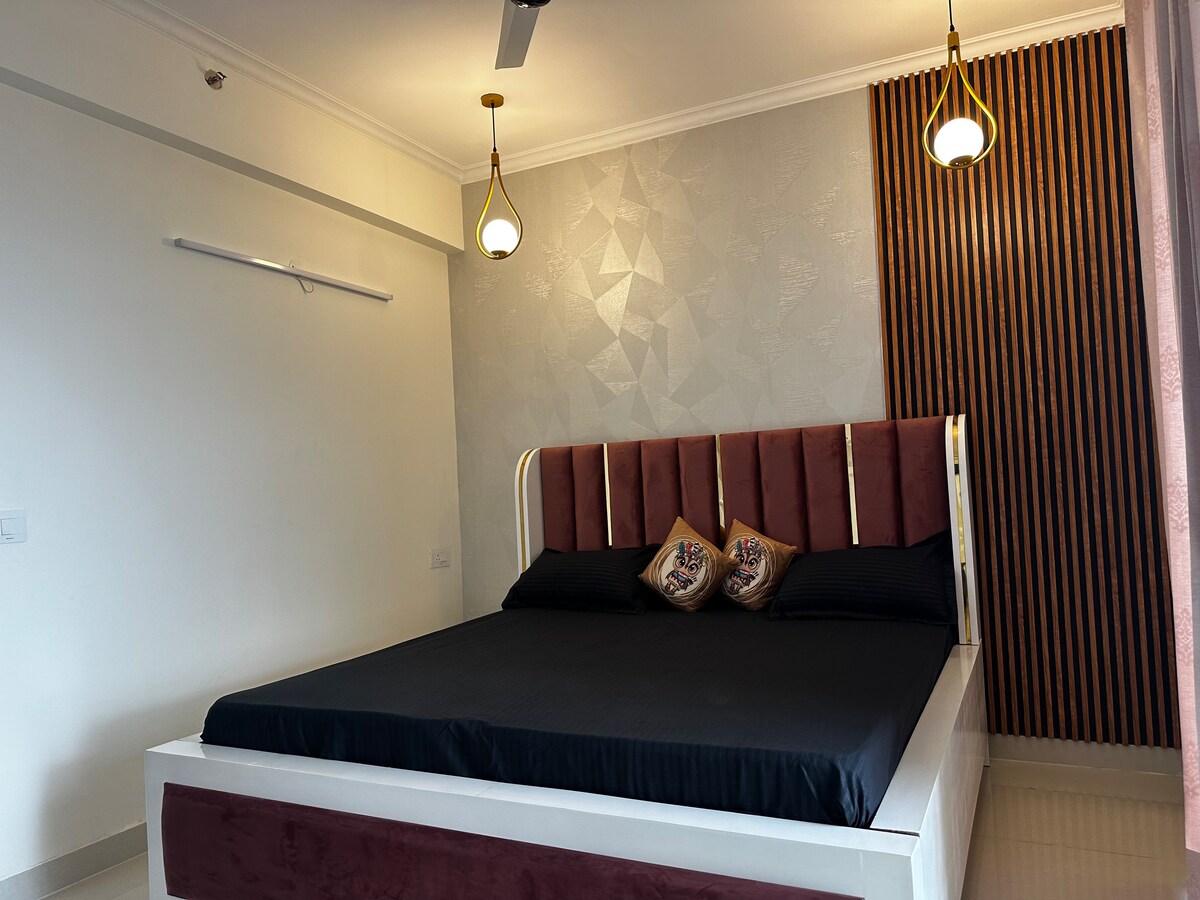 A spacious bedroom features a large bed with a dark bedding set and decorative pillows. Wall-mounted lighting fixtures and a textural accent wall enhance the room's modern design. Natural light enters through a nearby window, illuminating the clean lines and minimalist decor.