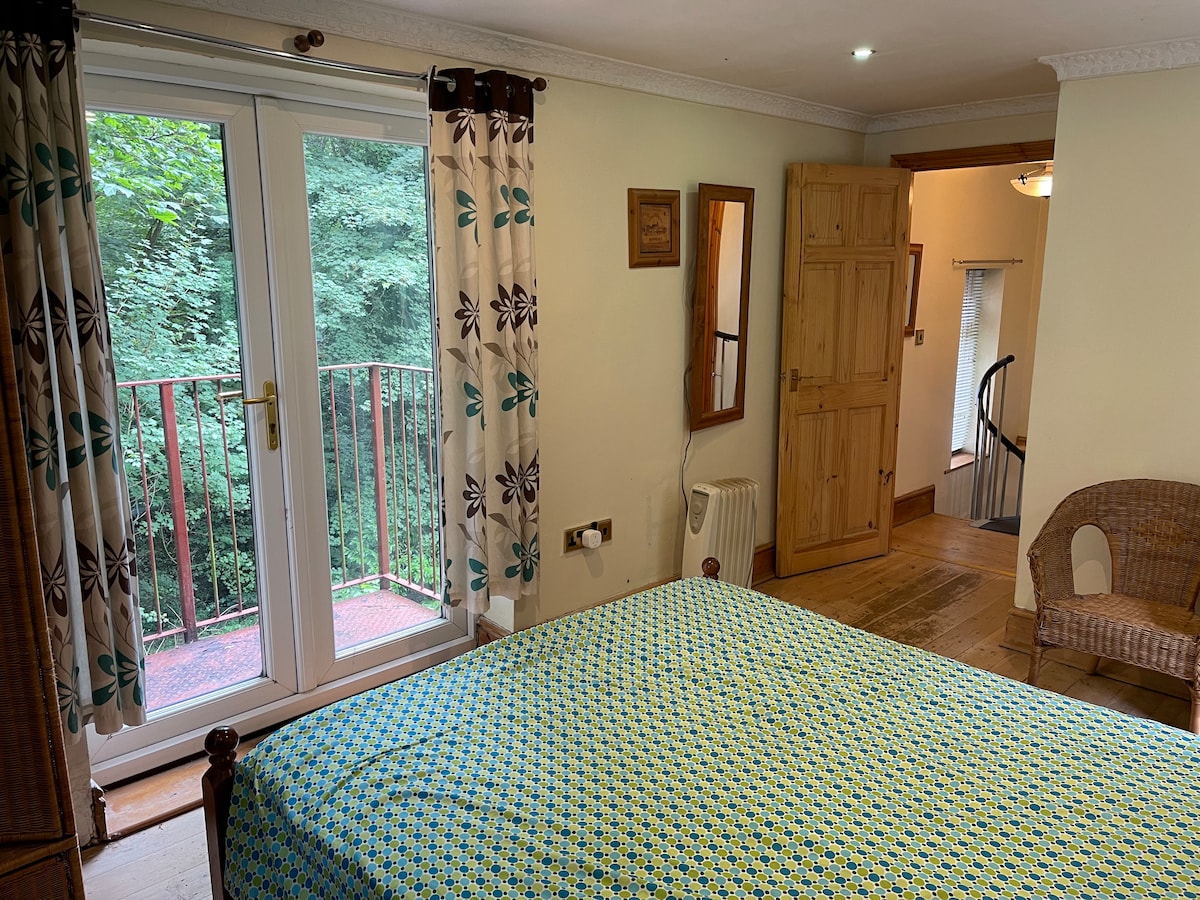 A spacious bedroom features a colorful patterned bedspread complemented by natural wood furnishings. French doors open to a balcony, inviting natural light and views of greenery. A woven chair is positioned nearby, with a mirror reflecting the cozy interior.