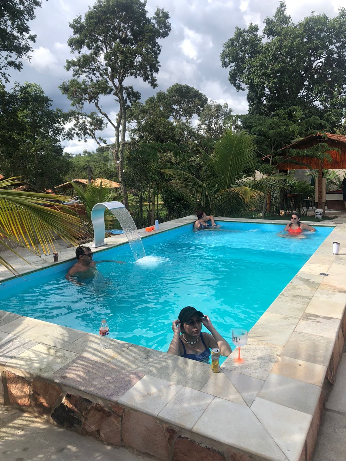 A serene swimming pool is surrounded by lush greenery, featuring a gentle waterfall. Guests are seen enjoying the refreshing water, while palm trees offer shade. A gazebo is visible in the background, enhancing the tranquil atmosphere of the outdoor space.