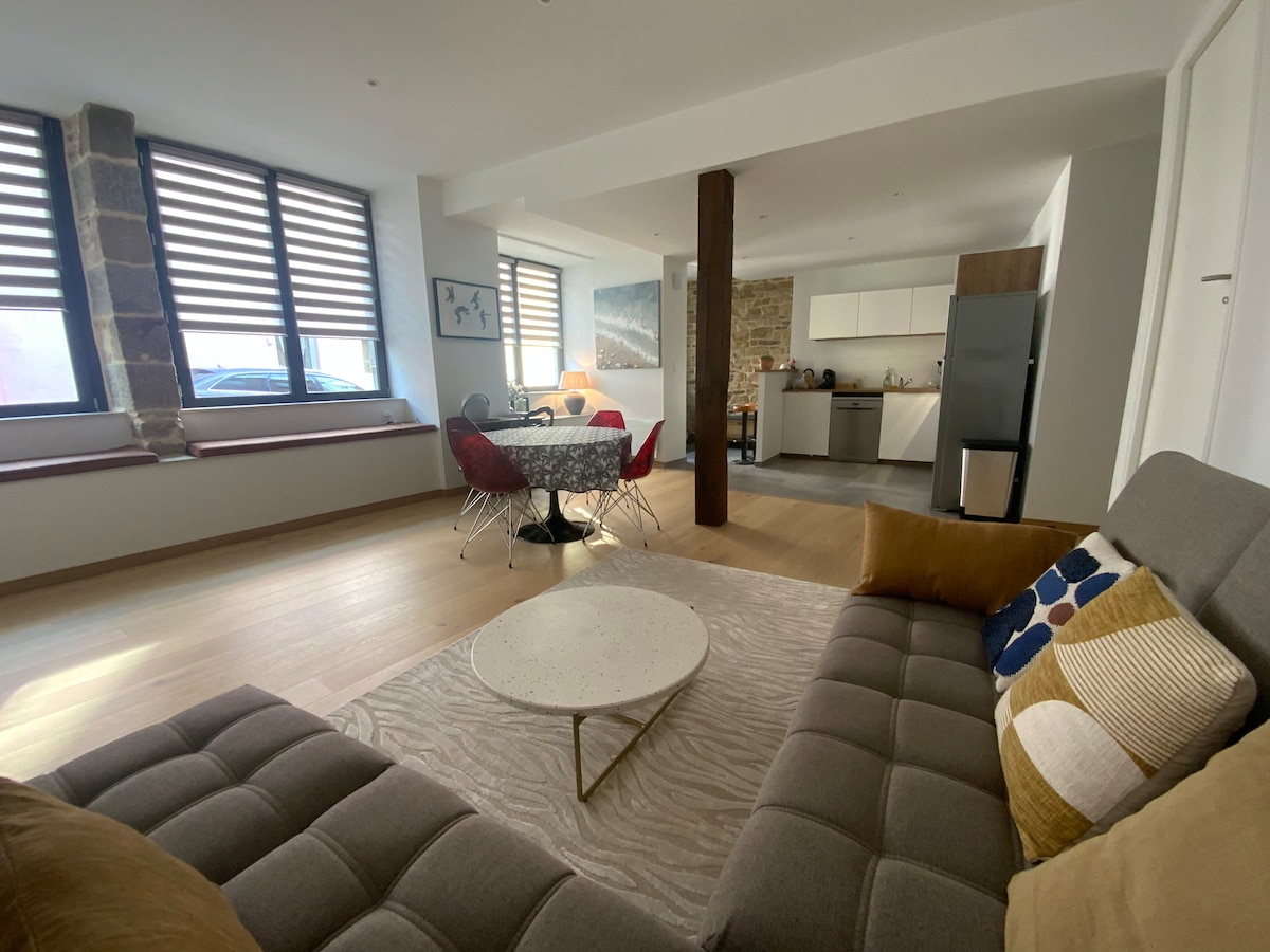 A spacious living area is shown, featuring a comfortable gray sofa with decorative pillows. A round table with red chairs is positioned next to the kitchen, which includes modern appliances. Natural light filters through window blinds, illuminating the wood flooring.