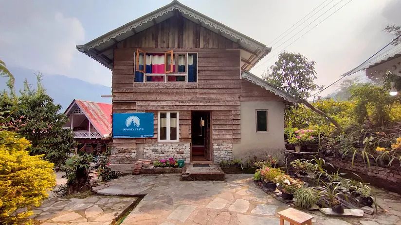 A charming two-story home displays a wood and stone exterior, framed by lush greenery and flower beds. A blue sign marks the entrance, while colorful fabrics hang from the upper windows, adding a touch of warmth to the inviting façade.