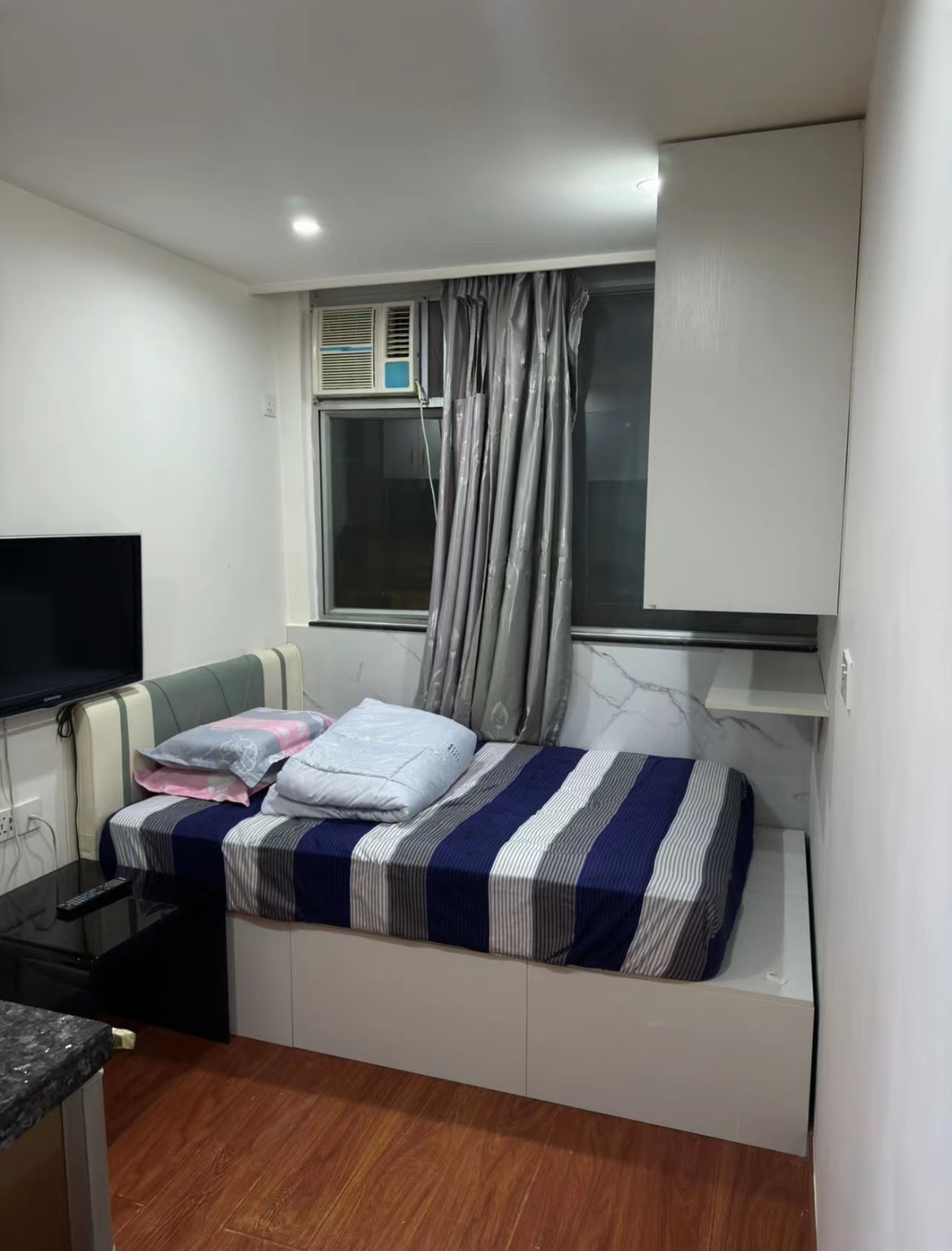 A compact bedroom is presented with a spacious bed featuring a striped blue and white cover. Pillows are neatly arranged on the bed. A television is mounted on the wall opposite the bed, and a window with light curtains provides natural light.