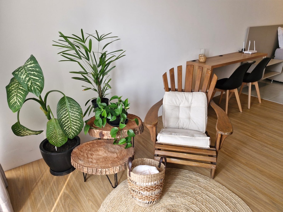 A comfortable wooden armchair is positioned beside two natural wood side tables, adorned with greenery from potted plants. A woven basket adds texture to the space, which features a light wooden floor. A spacious work desk and dining chairs are visible in the background.