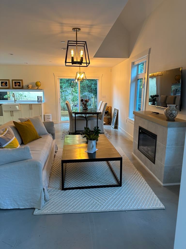 The living area features a comfortable couch with decorative pillows, facing a sleek coffee table on a patterned rug. A modern fireplace adds warmth, while a dining table is set beside a large window that allows natural light to illuminate the space.