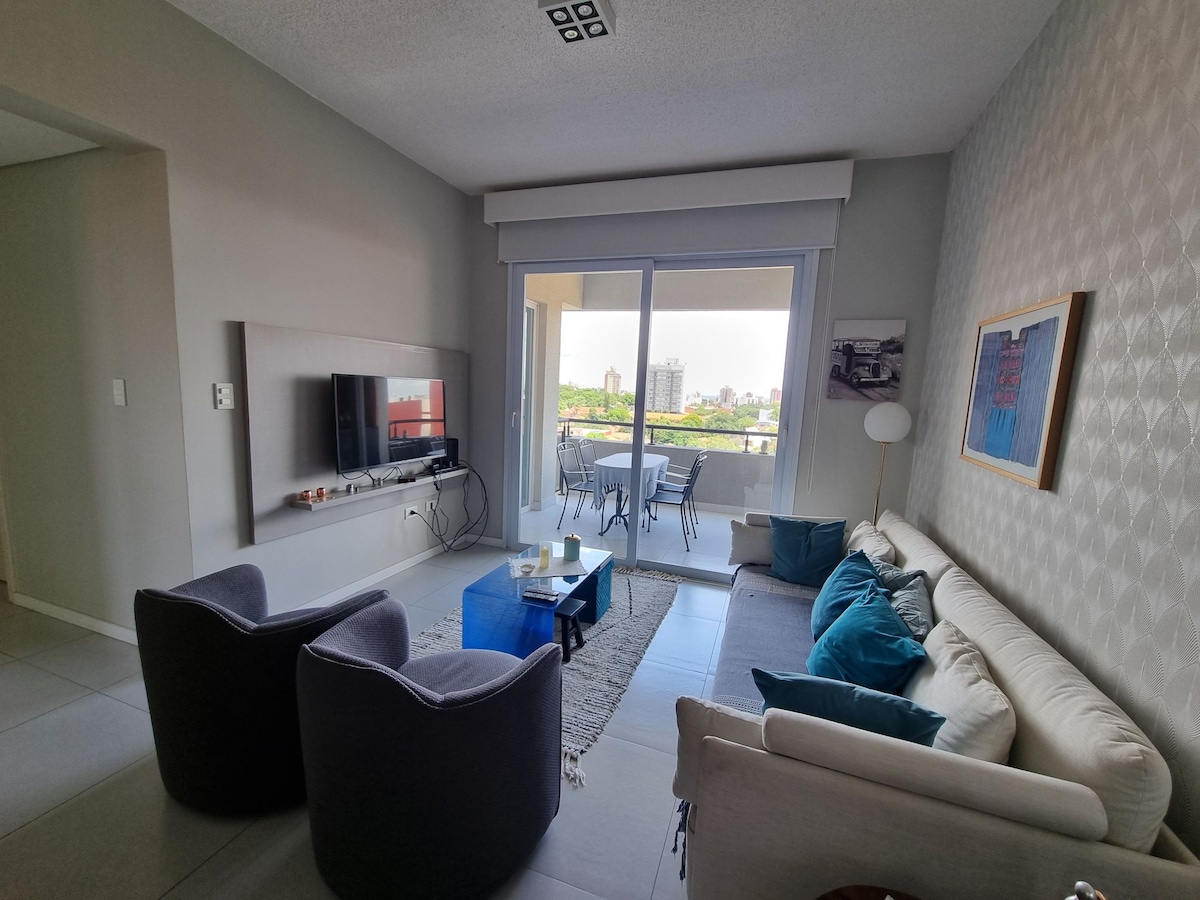 A modern living area is featured, furnished with a comfortable sofa adorned with cushions and two stylish armchairs. A glass coffee table rests on a soft rug, while large windows provide natural light and a view of the outdoor balcony with seating.