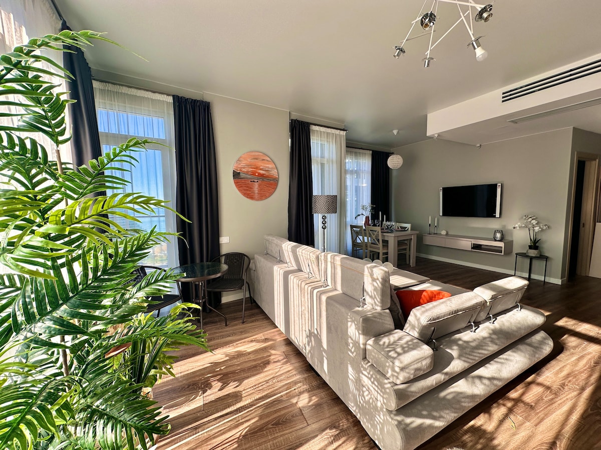 A spacious living area features large windows allowing natural light to fill the room. A comfortable sofa is positioned in the center, complemented by a dining table set for four. Decorative plants and modern art pieces add subtle touches to the inviting atmosphere.