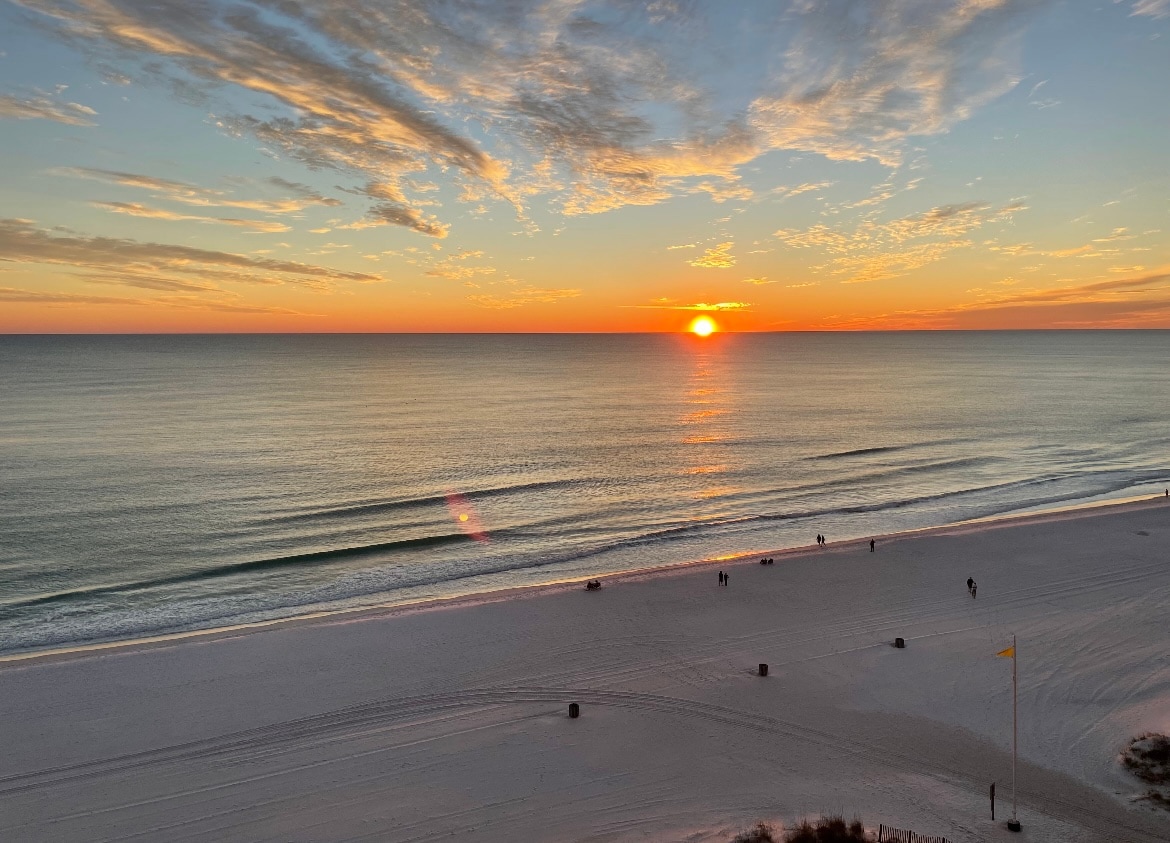 A serene beach landscape captures the setting sun, casting warm colors across the sky. Gentle waves lap at the shoreline, where people stroll along the sandy beach. Silhouette structures may be seen in the foreground, enhancing the tranquil ambiance of the scene.