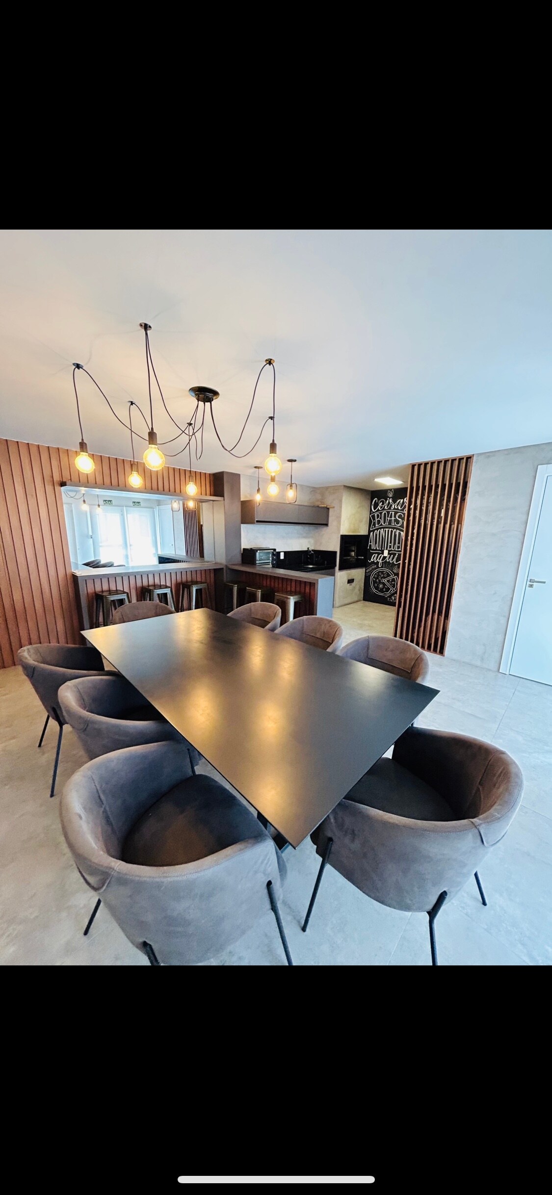 A modern dining area features a large black table surrounded by several plush, rounded chairs. Soft lighting is provided by a unique chandelier above, while wooden paneling adds warmth to the space. A glimpse of the kitchen is visible in the background.