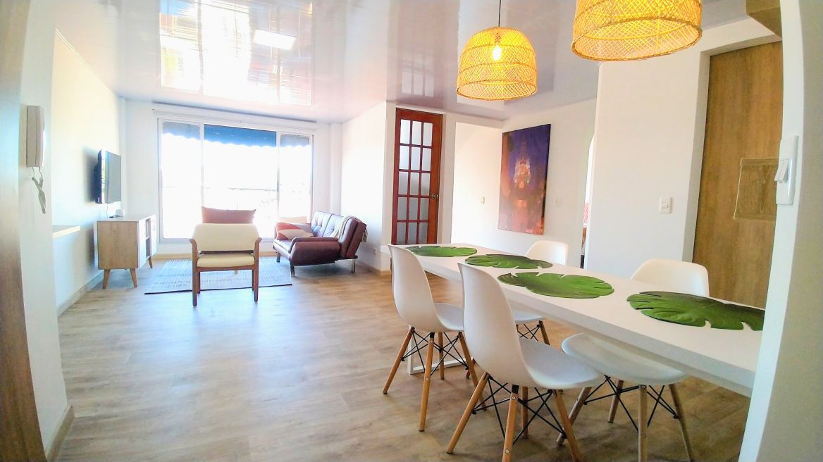 A spacious living area is presented, featuring a dining table with white chairs adorned with green placemats. A brown sofa and a comfortable chair are positioned for relaxation. Two pendant lights provide illumination, while large windows offer natural light and a serene view.