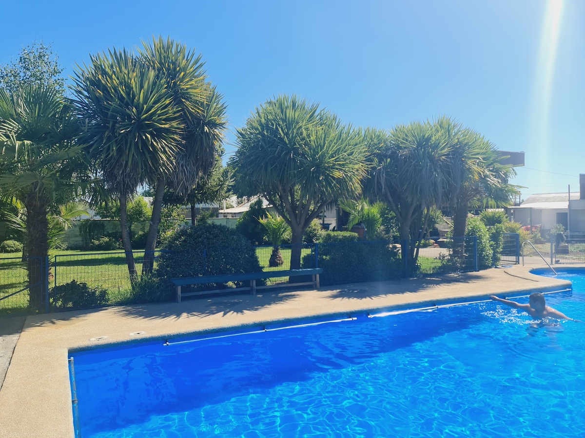A large swimming pool is surrounded by tropical palm trees and well-maintained greenery. A sunlit area is visible, providing space for relaxation. The tranquil ambiance is enhanced by the clear blue water, reflecting the bright sky above.