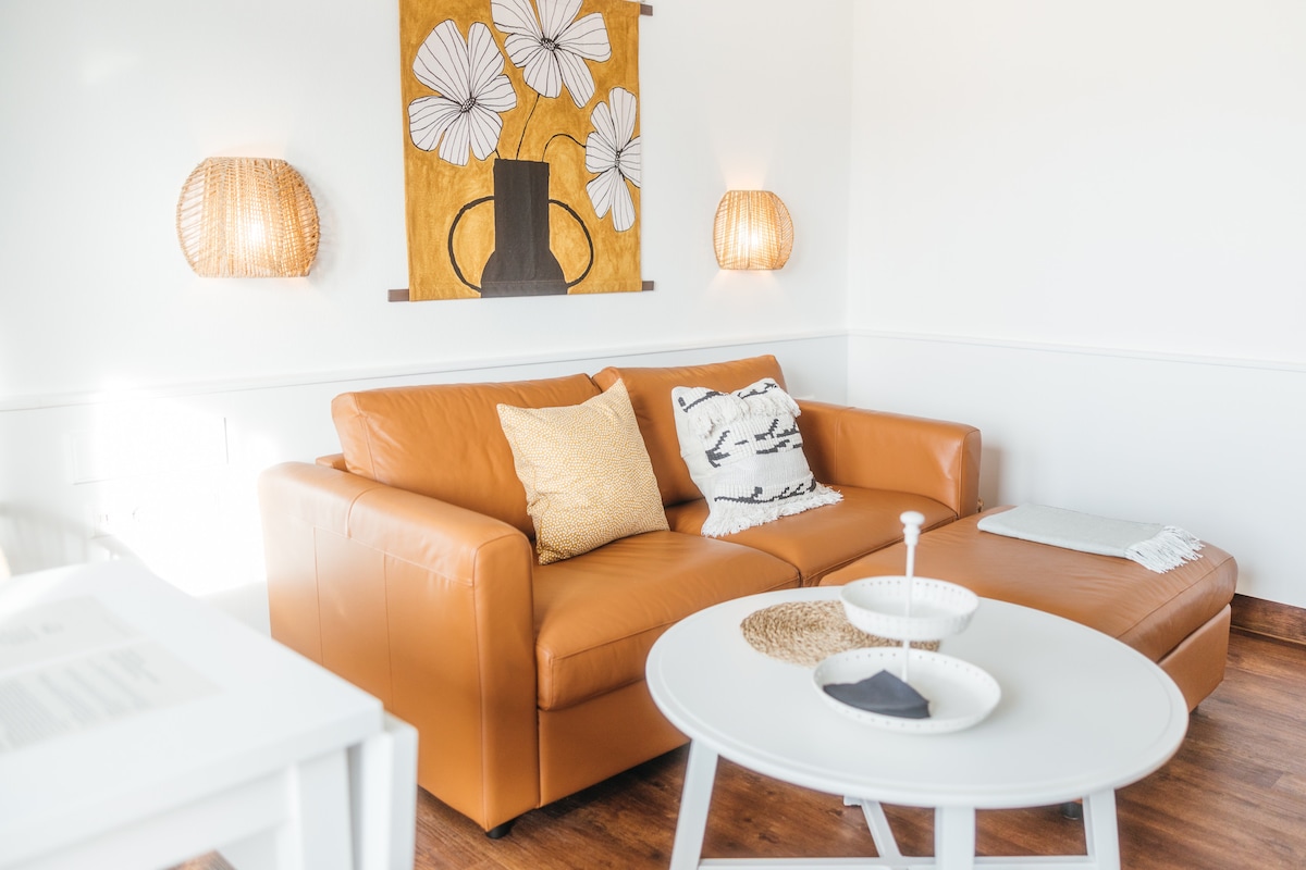 A welcoming living area features a warm leather sofa adorned with decorative pillows and a light throw. Soft lighting is provided by wall sconces, and a simple round table with decorative elements complements the design. A vibrant wall art piece adds a touch of color.