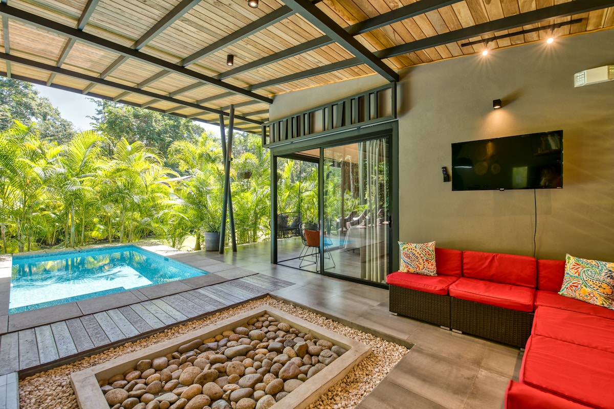 An outdoor living space features a cozy sectional sofa with vibrant red cushions, facing a sleek pool. Large glass doors provide views of lush greenery surrounding the area. A stone fire pit with smooth pebbles sits on a wooden deck, adding to the inviting atmosphere.