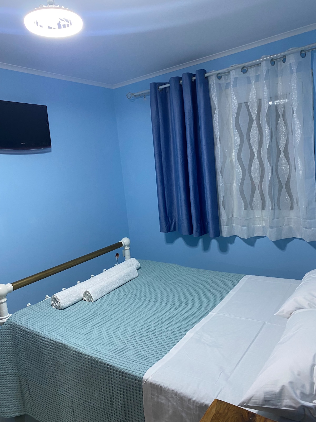 A cozy bedroom features soft blue walls, a double bed with a white and blue bedding set, and two rolled towels placed neatly at the foot. A small television is mounted on the wall, while sheer and dark blue curtains frame the window, allowing natural light to filter in.