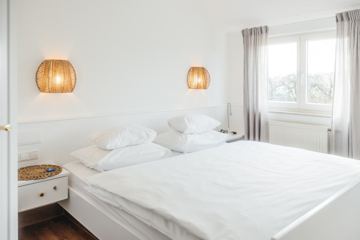 A bright bedroom features a spacious bed with white linens and multiple pillows. Two woven light fixtures on the wall provide soft lighting. A large window offers a view of the outdoors, framed by light, flowing curtains.