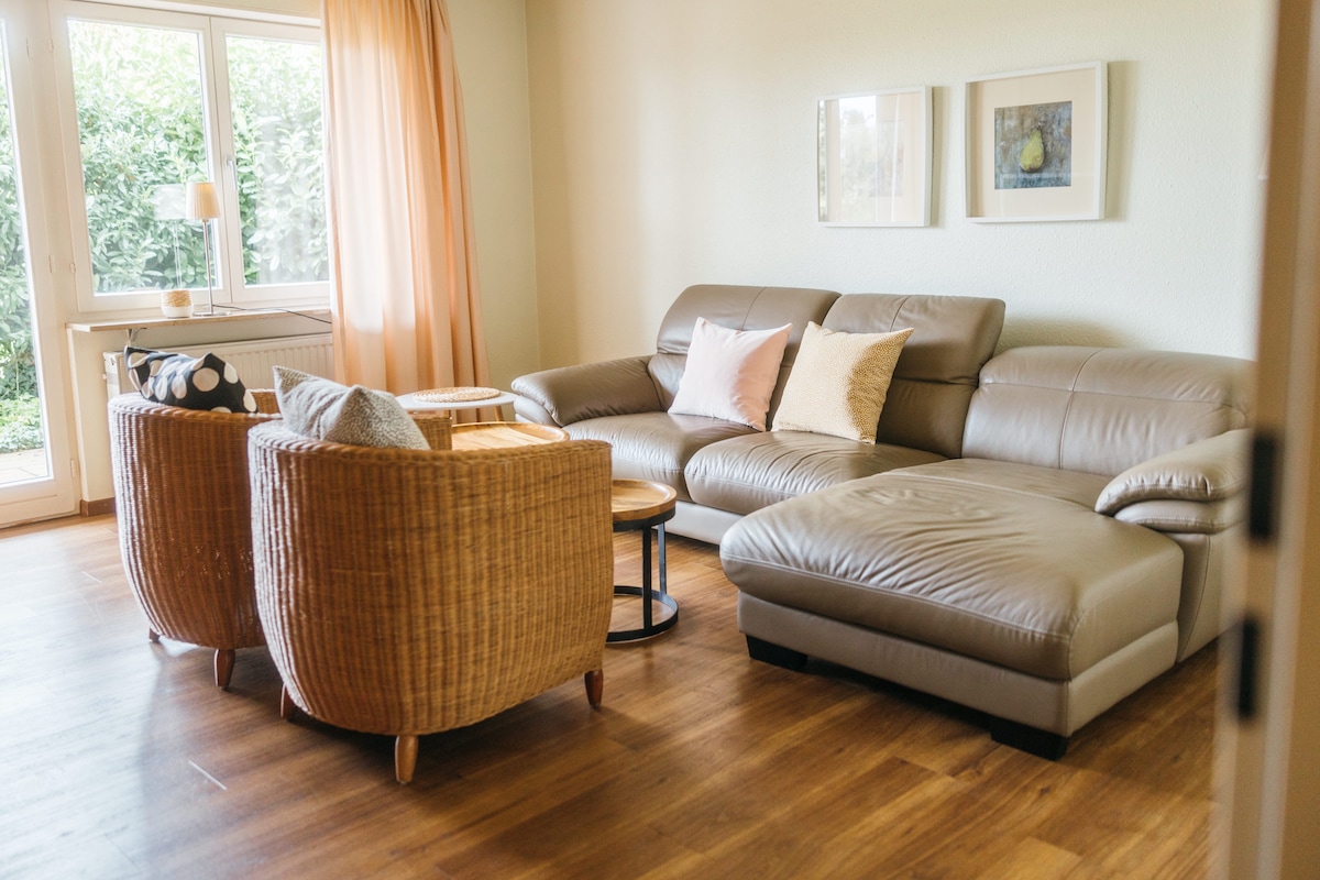A light-filled living room features a comfortable leather sectional sofa and two woven armchairs positioned around a small wooden table. Soft cushions add texture to the seating area. Natural light streams in through a large window dressed with sheer curtains, bringing warmth to the space.