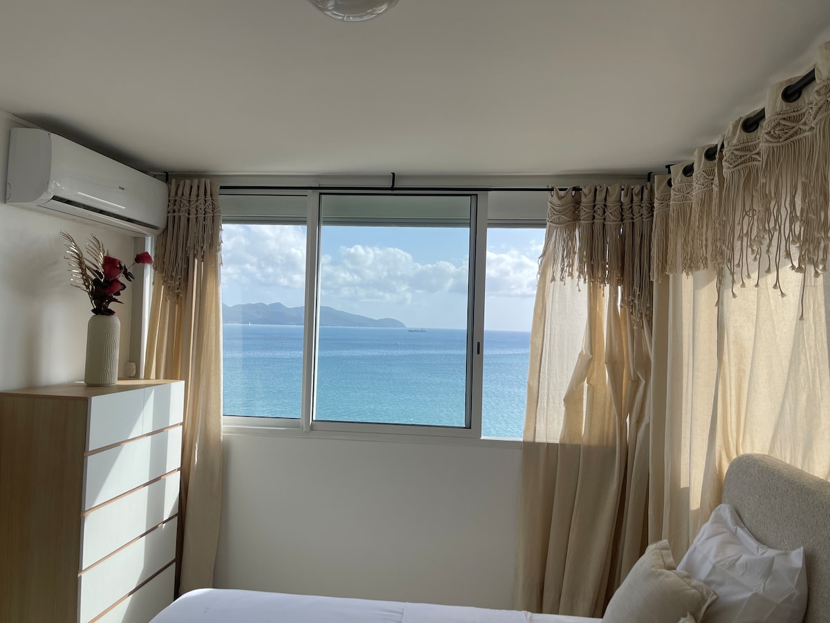 A bright bedroom features a spacious bed with light-colored linens. Large windows provide a view of the sea, allowing natural light to fill the space. Sheer curtains frame the windows, and a wooden dresser is positioned against the wall, adding to the serene atmosphere.