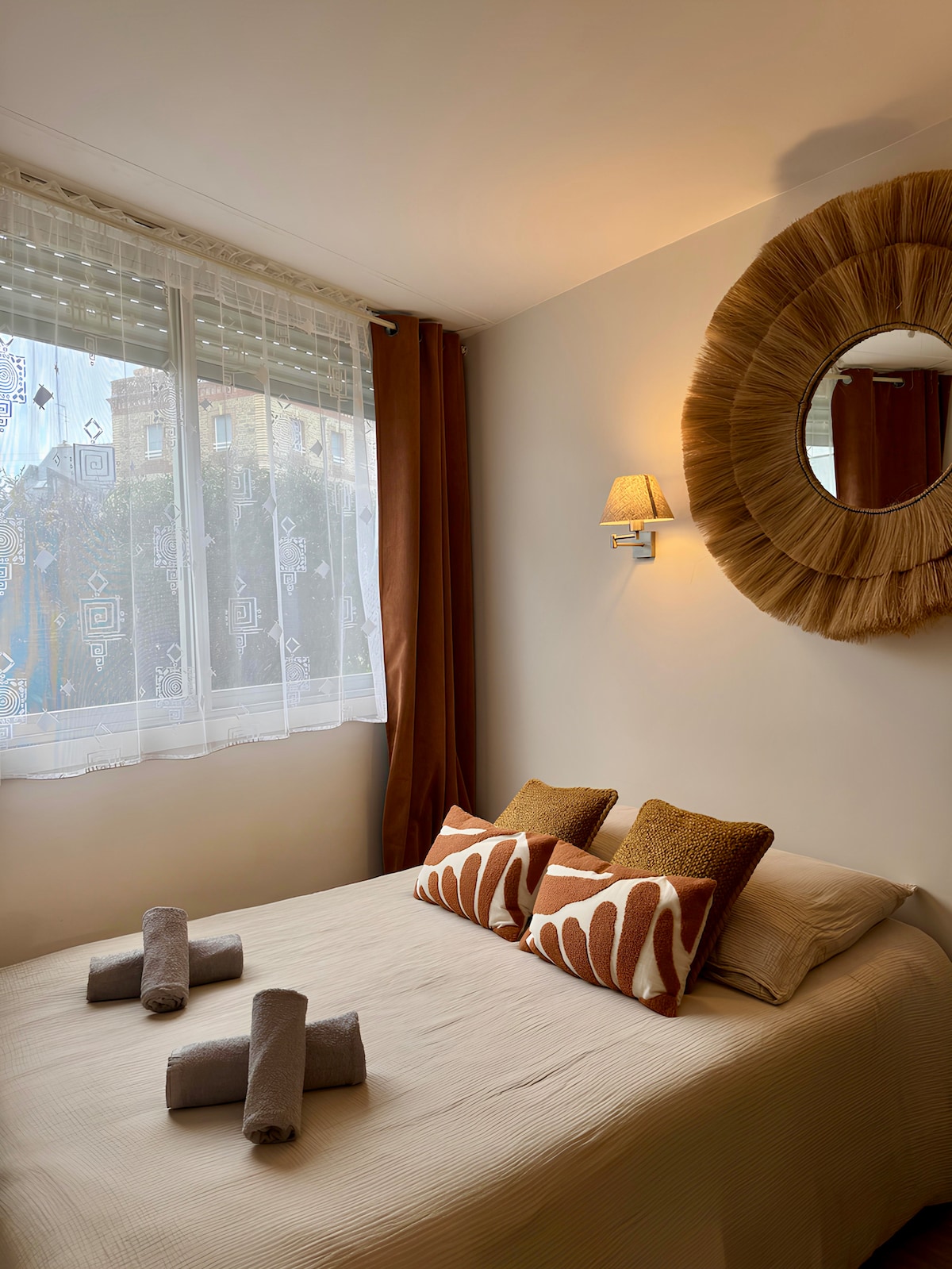 A cozy sleeping area is presented with a spacious bed adorned with neutral linens. Two decorative pillows and rolled towels are neatly arranged on the bed. Soft lighting emanates from a wall-mounted lamp, while natural light filters through sheer curtains, creating a serene ambiance.