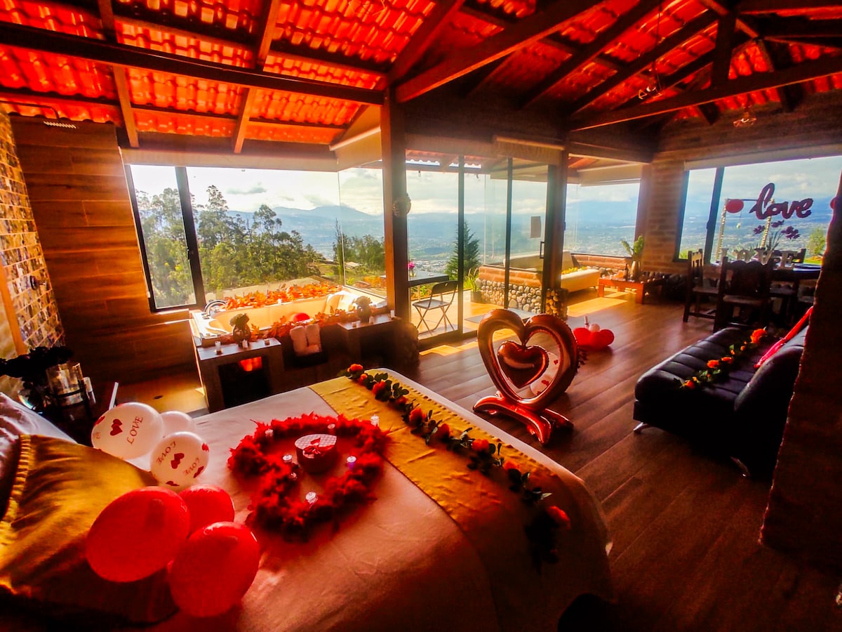 The open interior features a spacious bed adorned with heart-shaped decorations. Large glass windows provide expansive views of the mountains and valley. A cozy seating area is positioned nearby, with additional decorations that enhance the romantic ambiance of the space.
