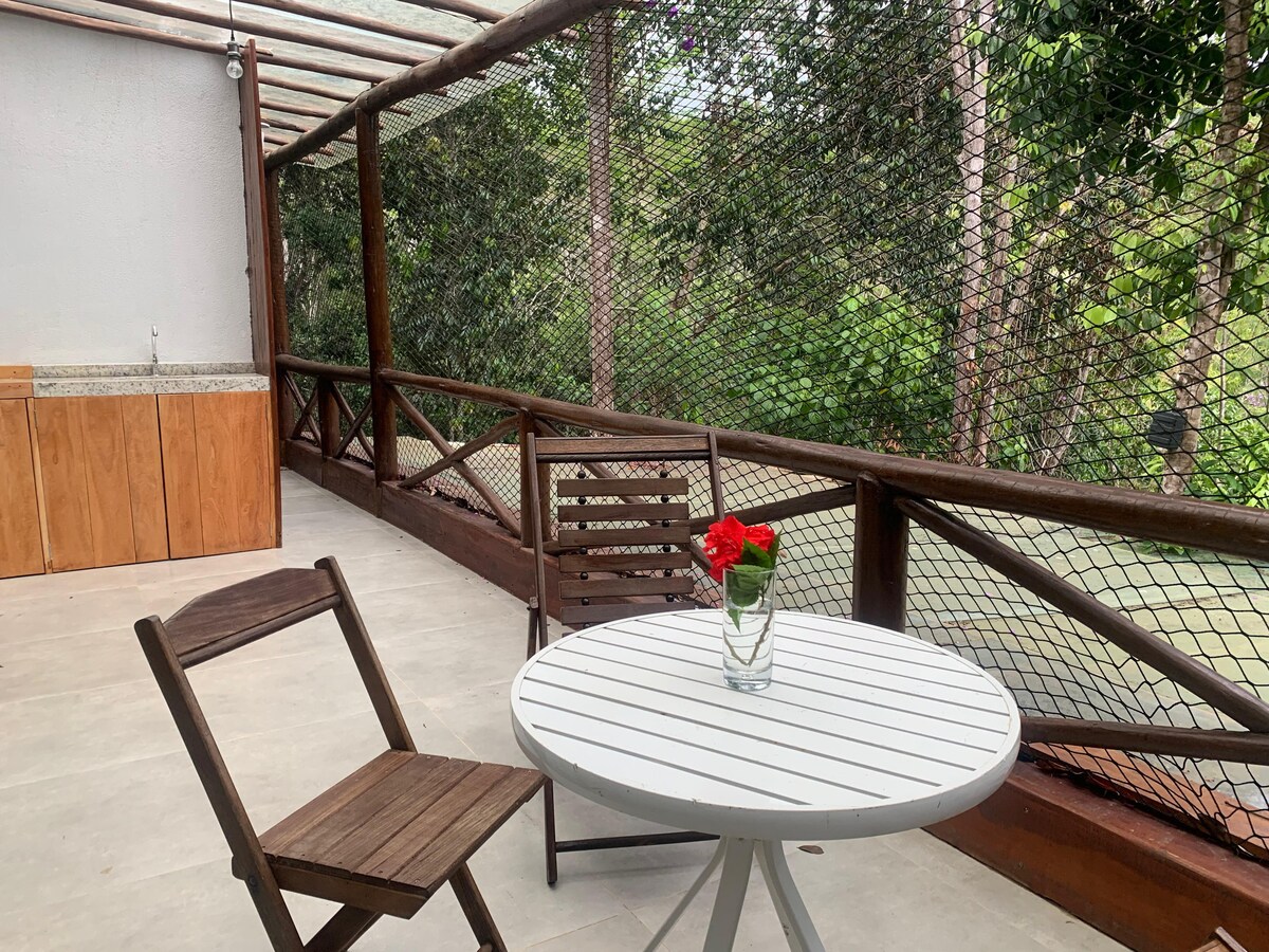 An outdoor seating area features a round white table accompanied by two wooden chairs. A small vase with vibrant red flowers adorns the table. Lush greenery is visible through the mesh fencing, enhancing the connection with nature.