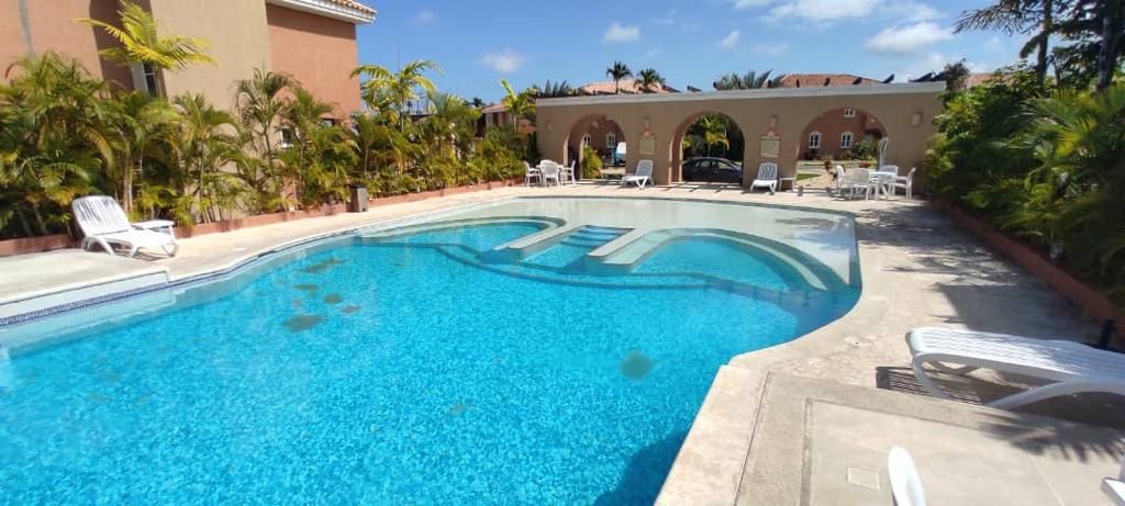 A spacious outdoor pool area is surrounded by lush greenery and palm trees. Lounge chairs are positioned around the pool, which features gently curved edges and steps leading into the water. Sunshine reflects off the clear blue water, creating an inviting atmosphere.