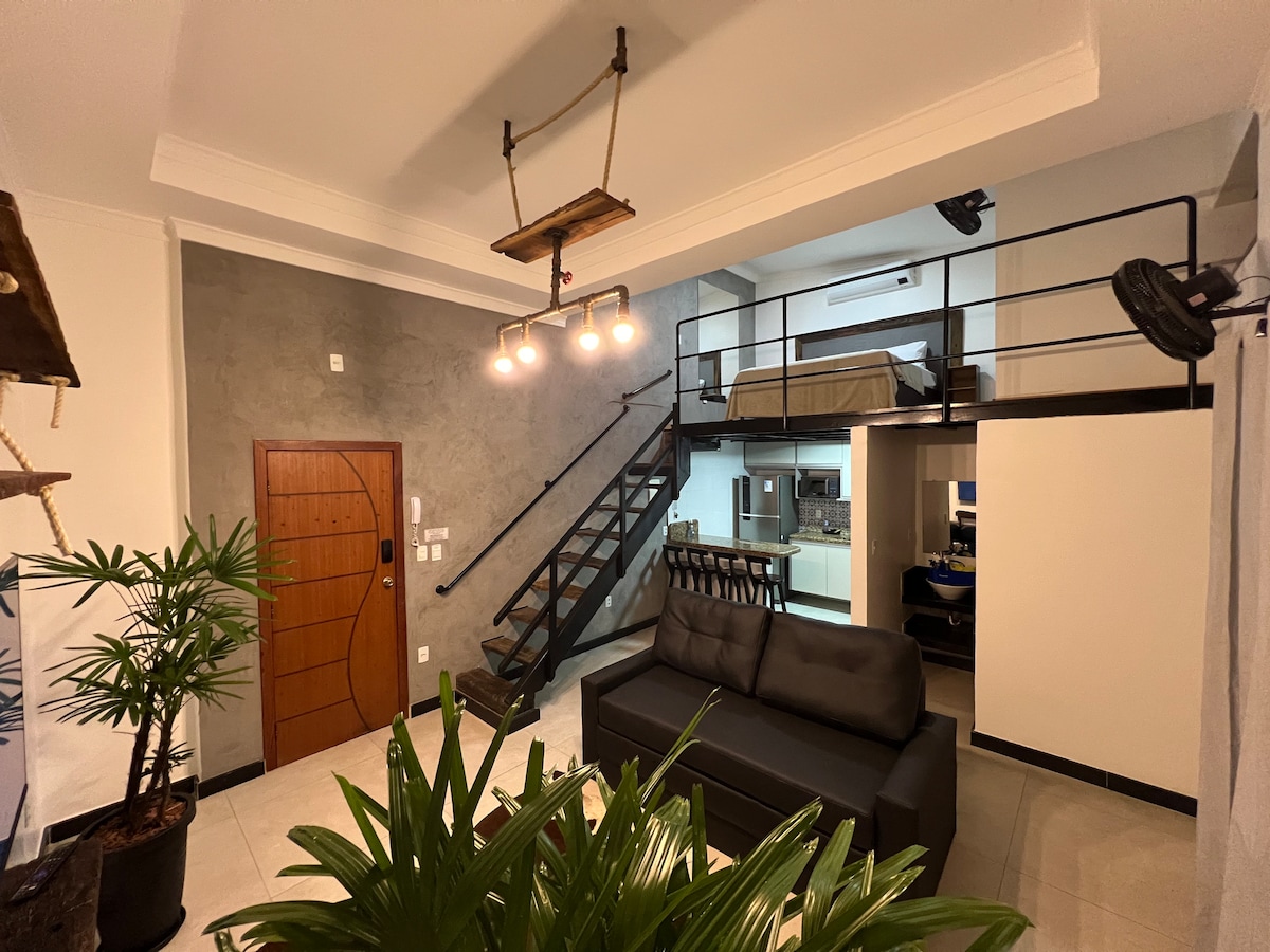 The open-plan living area features a comfortable sofa facing a set of stairs leading to a lofted bedroom. A dining table is positioned near the entrance, with light fixtures hanging above. Green plants add a touch of nature to the modern space.