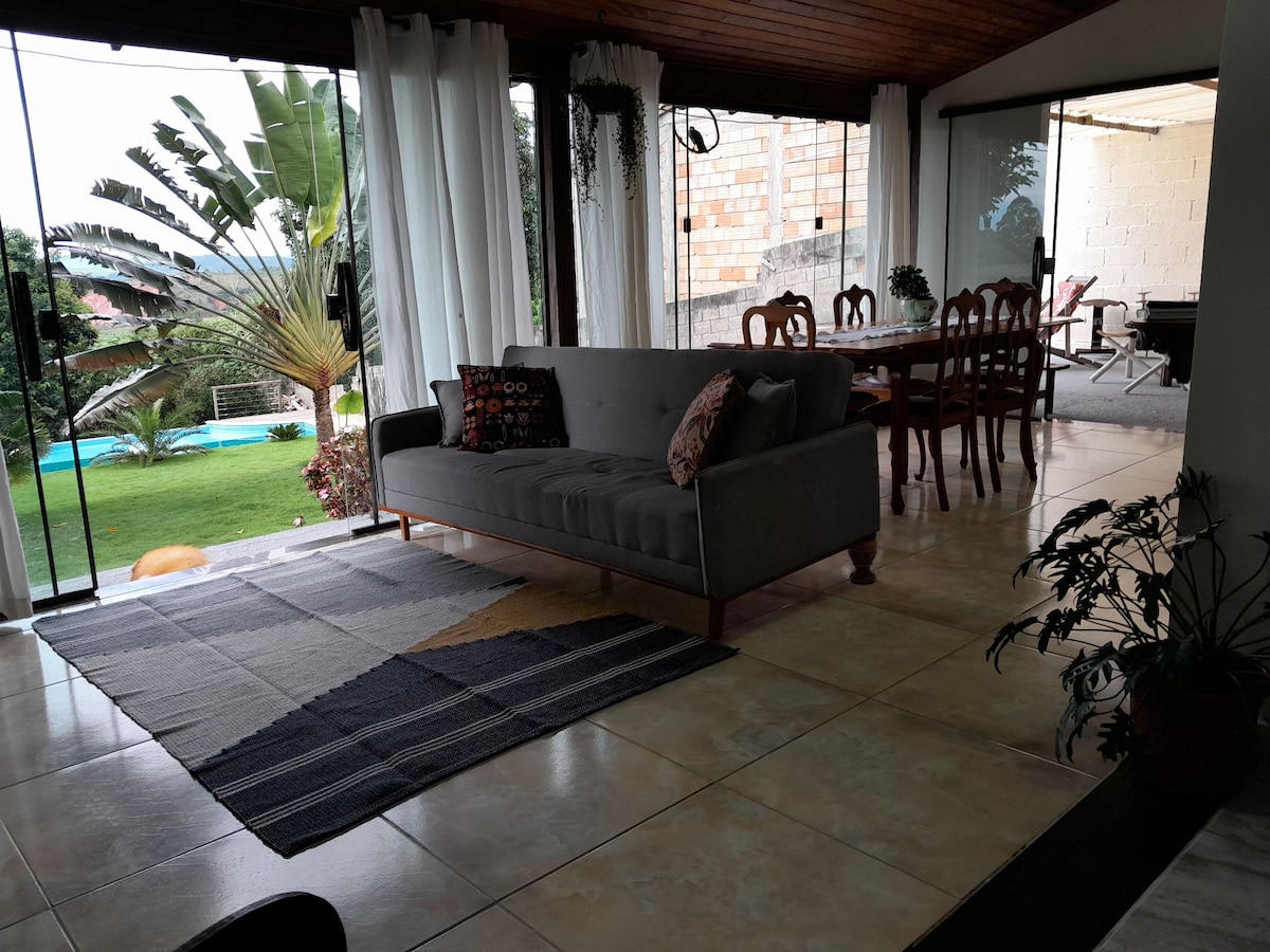 A spacious living area features a gray sofa adorned with decorative pillows, set against large glass doors that open to a lush garden. A dining table is positioned nearby, complemented by a patterned area rug and natural light flowing through sheer curtains.