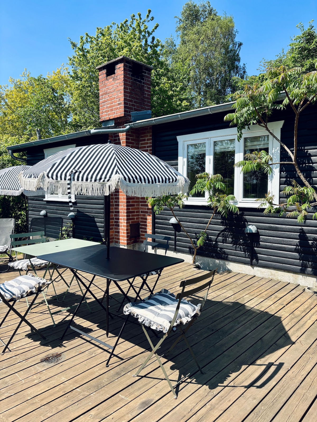 A wooden terrace is featured, showcasing a green table surrounded by four folding chairs. Two striped umbrellas provide shade, and the black wooden house stands in the background. Lush green trees and blue skies enhance the peaceful outdoor setting.