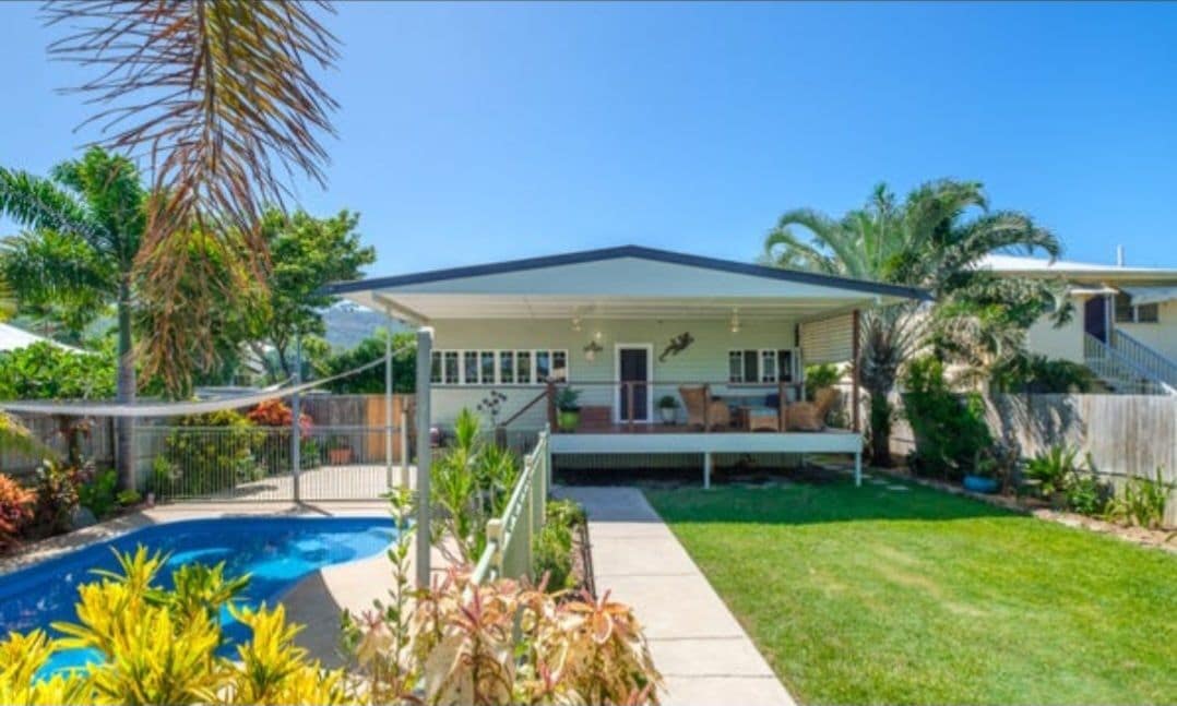 The exterior of the home is shown, featuring a covered front porch with seating and a well-kept lawn. A large pool is surrounded by a safety fence, and tropical plants frame the yard, creating a relaxed outdoor space. Bright sunshine enhances the inviting atmosphere.