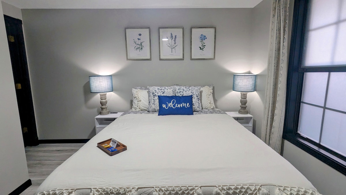 A spacious bedroom is presented with a large bed adorned in neutral tones, featuring decorative pillows and a welcoming message. Two bedside tables with lamps provide soft illumination. Floral artwork is displayed on the wall, enhancing the serene atmosphere of the room.