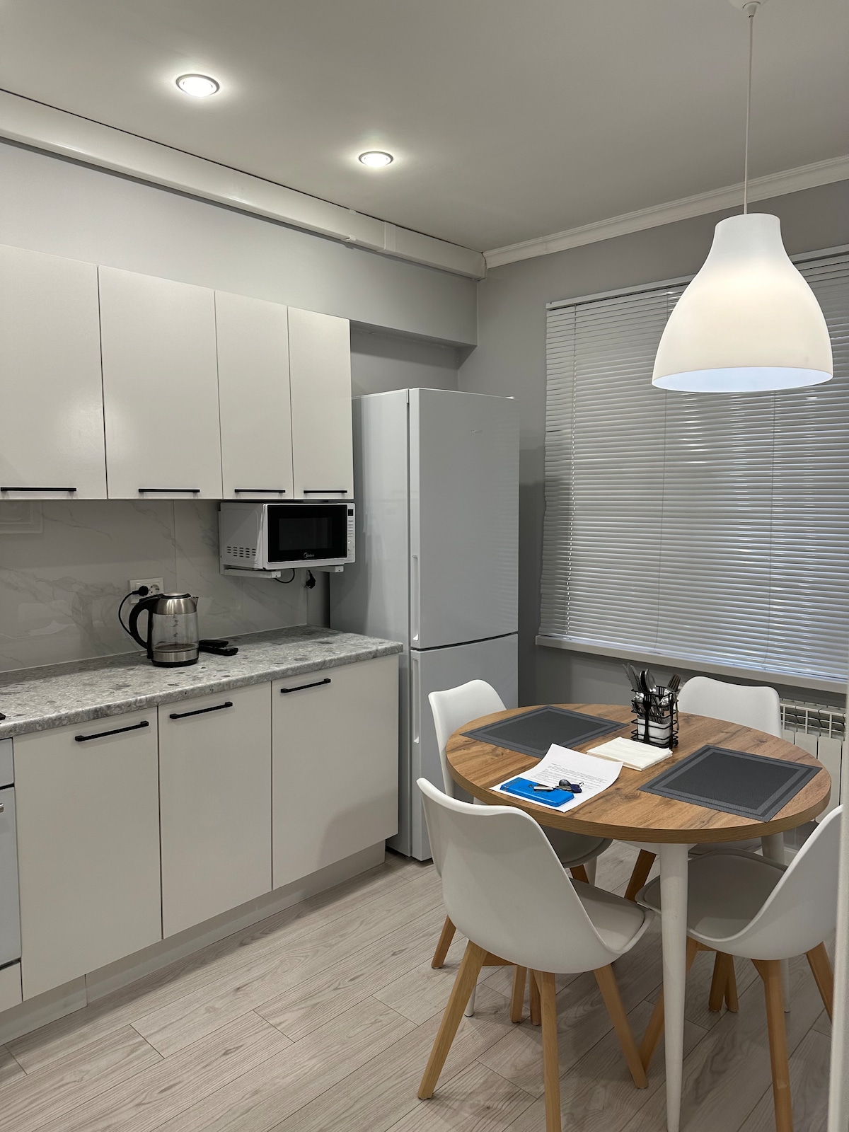 A modern kitchen area features sleek white cabinets and a light gray countertop. A circular wooden table with white chairs is set for dining. A refrigerator and a microwave are visible, along with a kettle on the counter. Natural light enters through the window, which is dressed with horizontal blinds.