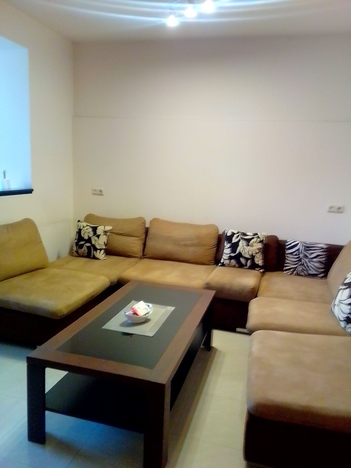 A cozy seating area features a sectional sofa with light brown upholstery and patterned throw pillows. A dark wood coffee table sits at the center, adorned with a decorative tray and small items. Soft overhead lighting enhances the inviting atmosphere.