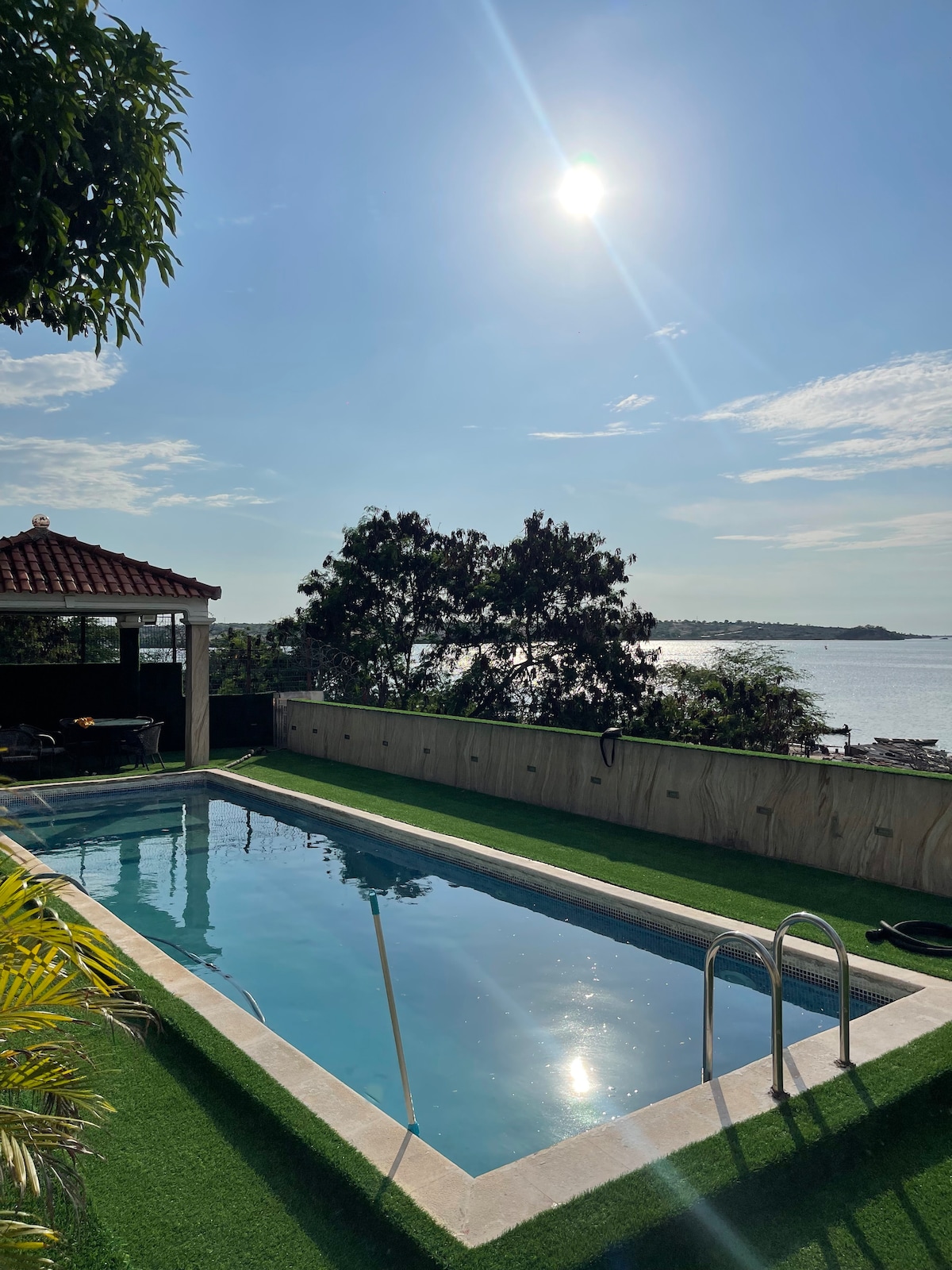 A serene pool is highlighted in a tranquil outdoor setting, reflecting the sunlight. Lush greenery surrounds the area, with a view of the ocean in the background. A shaded seating area is partially visible nearby, offering a space for relaxation.