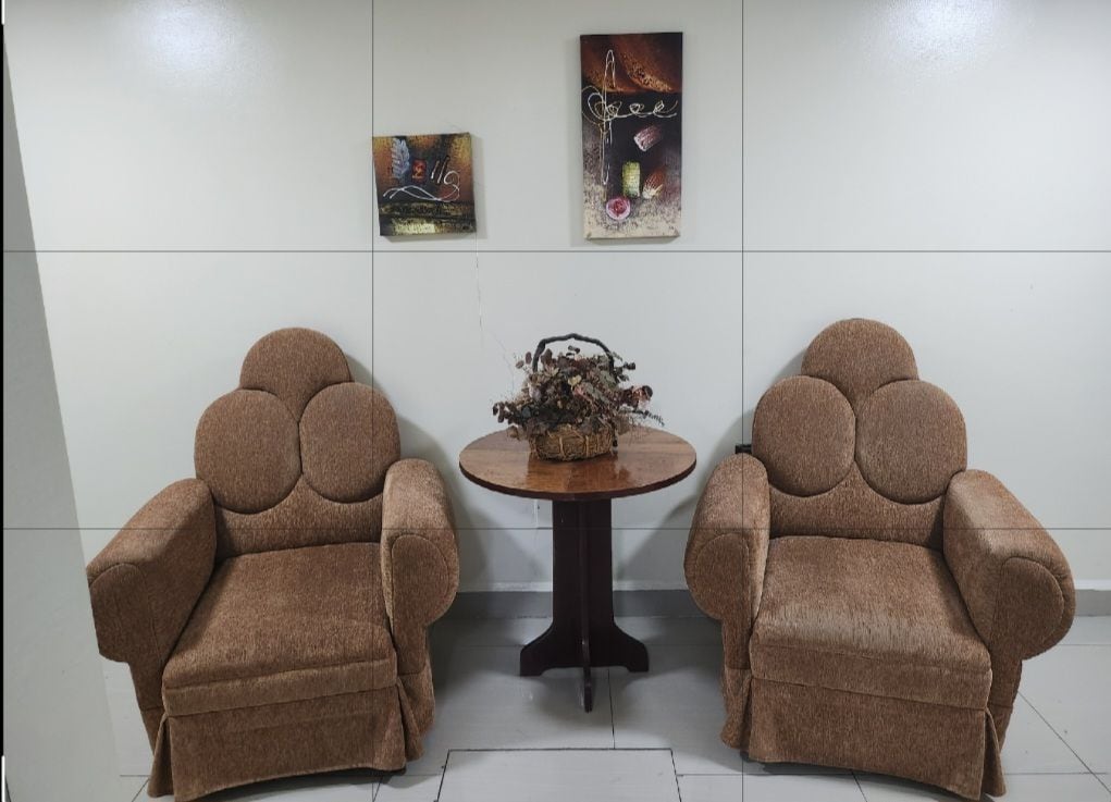 Two comfortable armchairs with a textured fabric are positioned on either side of a small round table. A decorative arrangement of dried flowers sits atop the table, flanked by two wall art pieces that add a touch of color to the neutral wall.