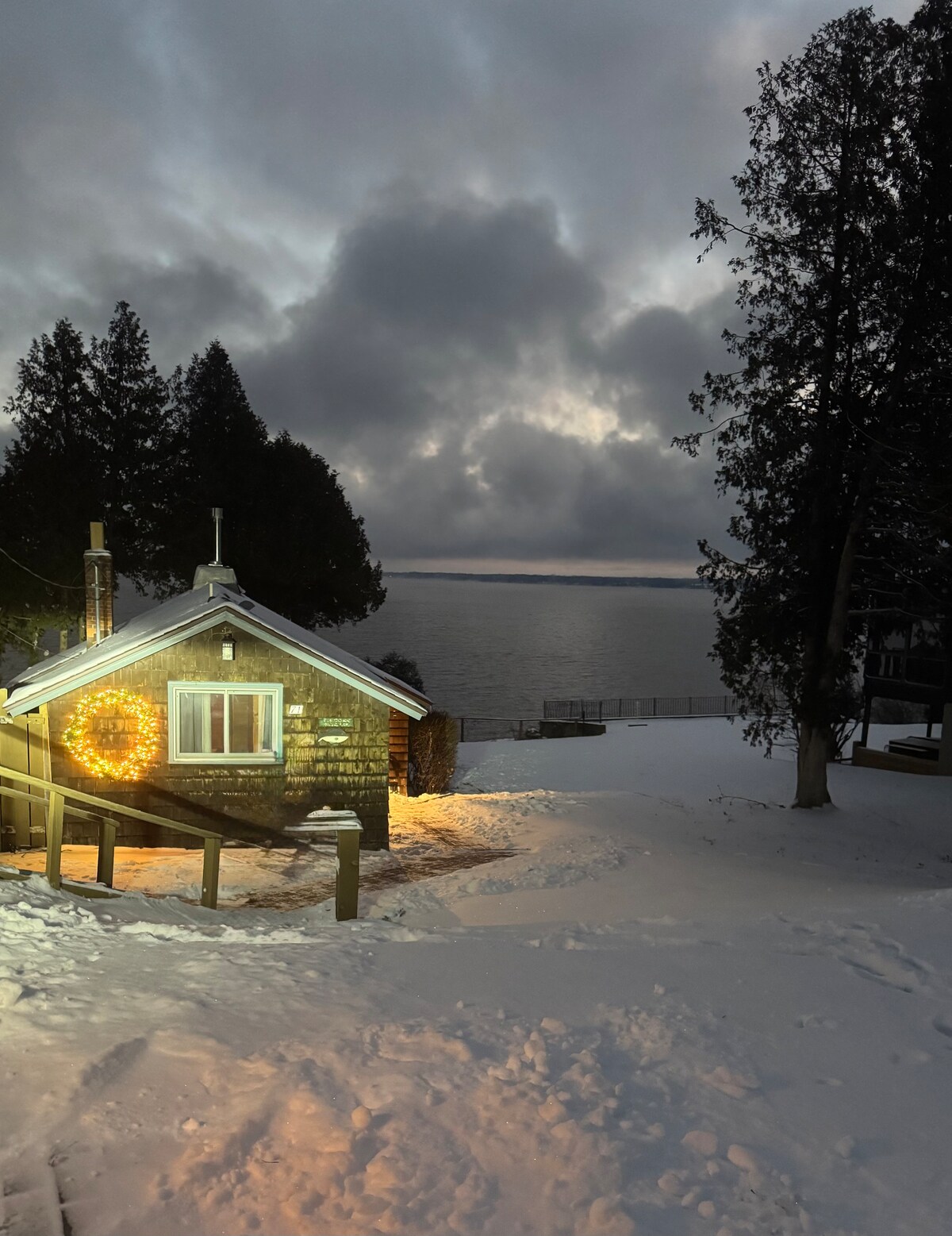 The cozy lakeside cottage is framed by snow-covered ground, with soft lighting illuminating the entrance. The lake glistens in the background under a moody sky, while tall trees stand nearby, enhancing the serene winter landscape.
