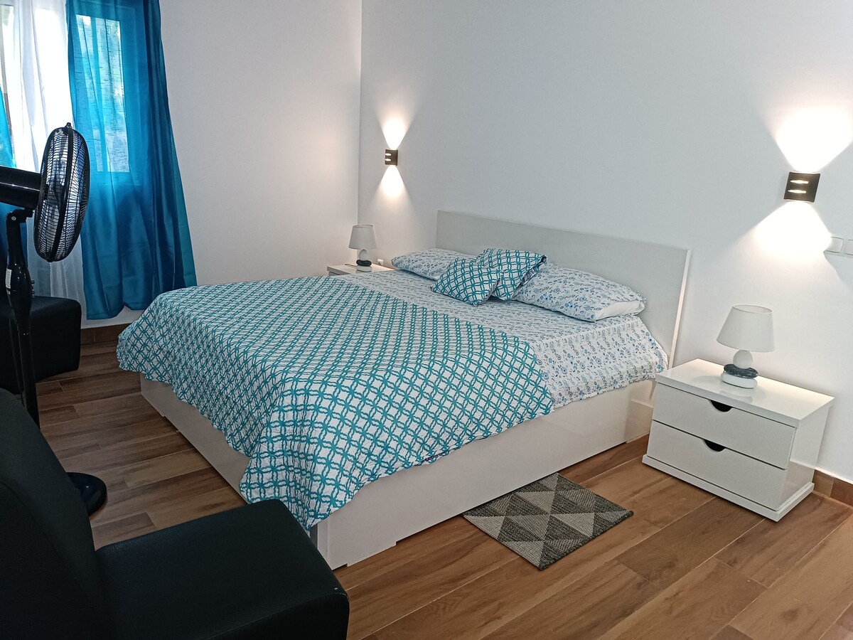 A well-lit sleeping area features a spacious bed dressed in a patterned blue and white quilt. Soft lighting is provided by bedside lamps on either side, and a fan is positioned nearby. Large curtains in vibrant blue frame the window, adding a touch of color.