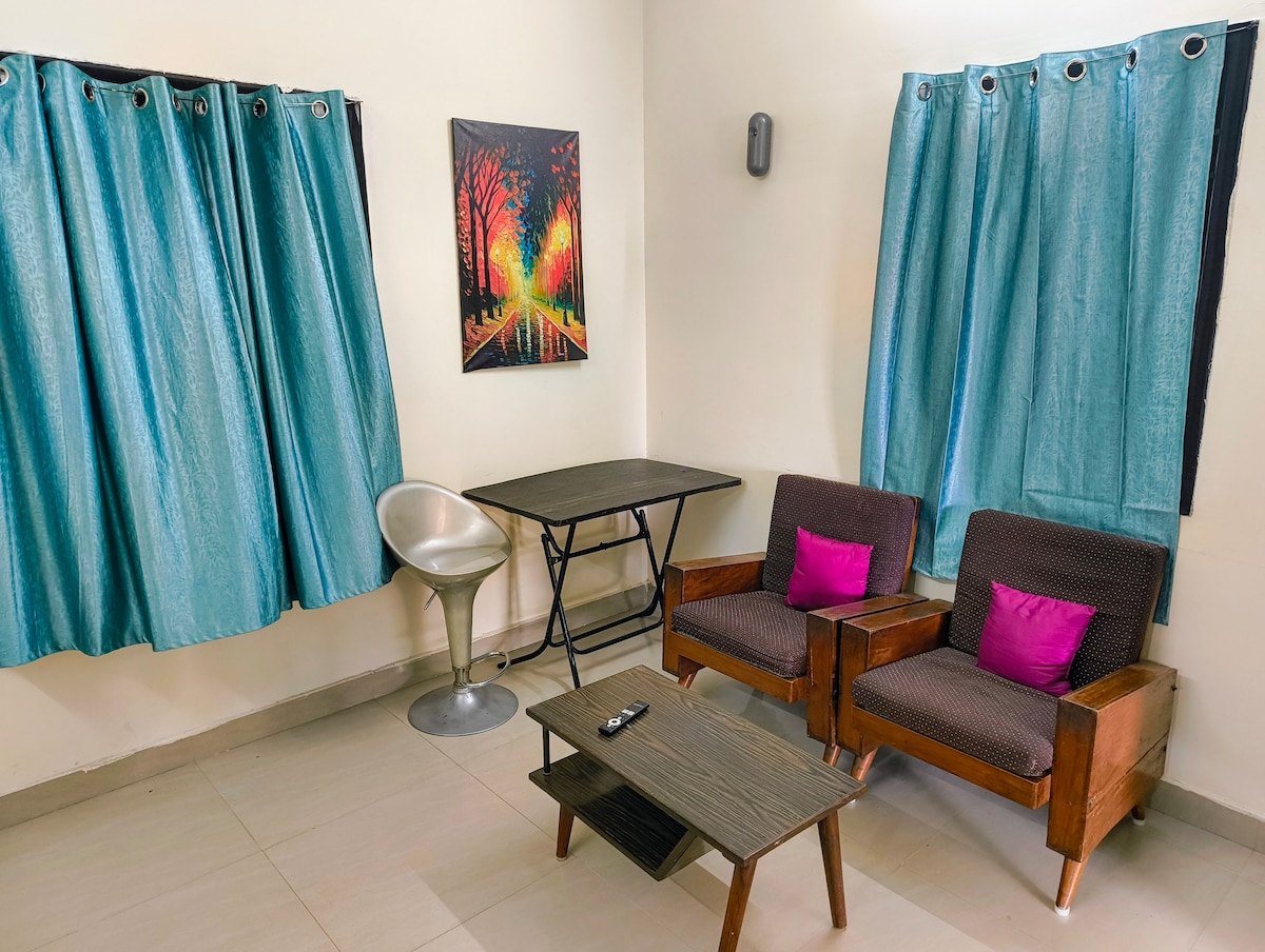 A cozy sitting area features two wooden chairs with purple cushions and a compact coffee table. A small, metallic chair complements the space. Bright turquoise curtains frame a window, while a colorful artwork of trees hangs on the wall, adding a vibrant touch to the room.