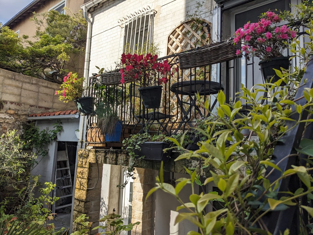 A charming balcony adorned with planters overflowing with vibrant flowers is visible. The exterior walls feature a whitewashed finish, complemented by wrought iron railings. Lush greenery surrounds the area, creating a serene setting that blends with the tranquil garden below.
