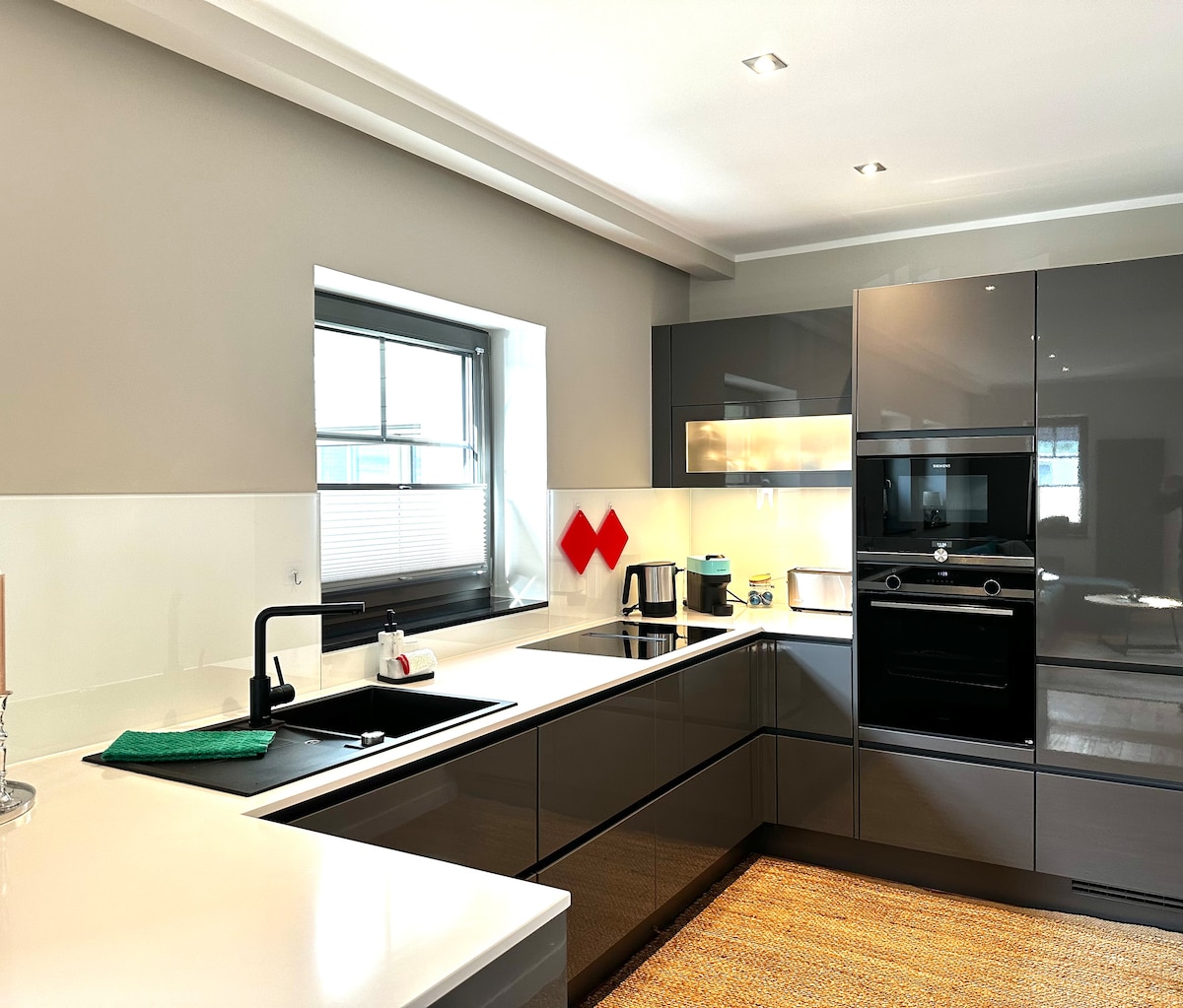 A modern kitchen is equipped with sleek grey cabinetry and a spacious white countertop. Stainless steel appliances, including an oven and stovetop, are visible. A window allows natural light to fill the space, creating a bright and functional cooking area.