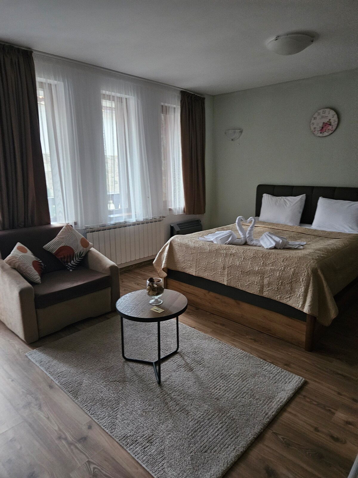 A well-lit room is furnished with a spacious bed topped with white linens. A comfortable armchair with decorative pillows is positioned next to a small table. Large windows with sheer curtains allow natural light to fill the space, enhancing its relaxed atmosphere.