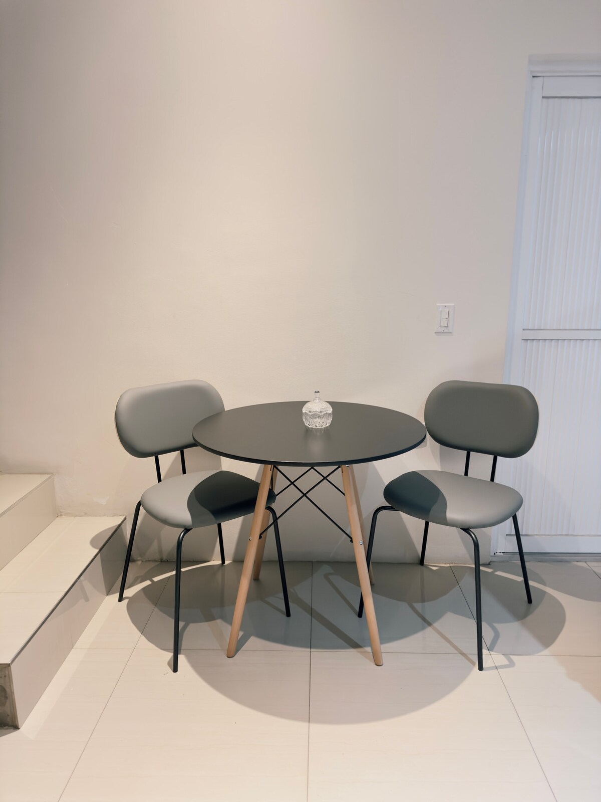 A small round black dining table is accompanied by two grey chairs, positioned against a neutral wall. A glass vase rests at the center of the table. Light-colored tiled flooring extends into the background, complemented by a white door.