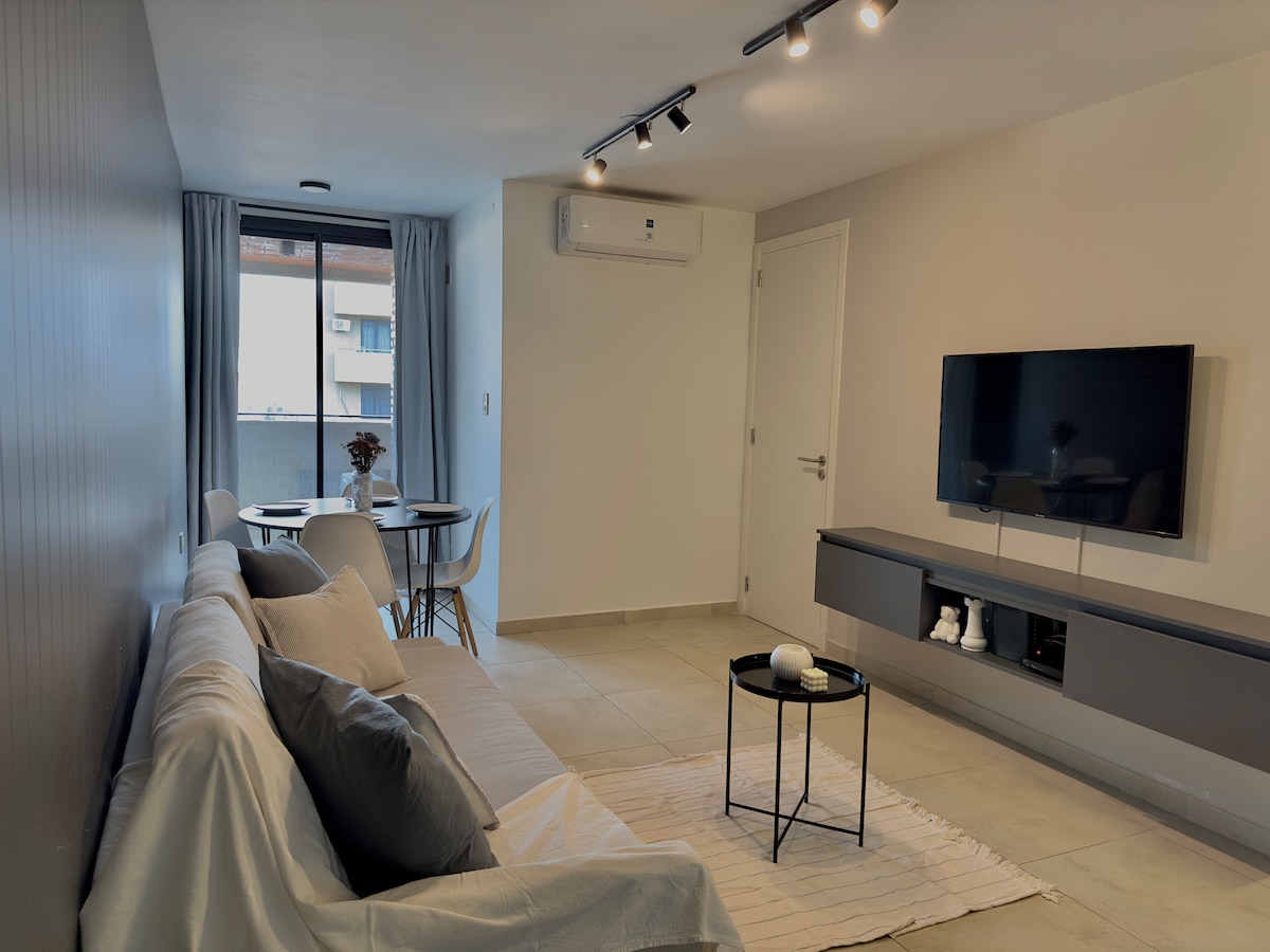 A modern living area features a light-colored sofa adorned with decorative pillows. A small black coffee table sits on a textured area rug, with a dining table and white chairs positioned nearby. Wall-mounted television and air conditioning unit are visible, accompanied by natural light from a window.