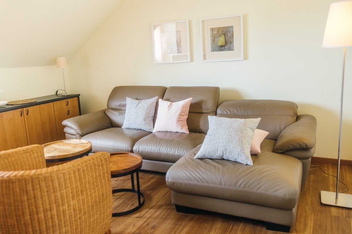 A comfortable living area is featured, showcasing a light-colored leather sectional sofa adorned with soft pillows in shades of pink and blue. Two circular wooden tables are positioned beside the sofa, while a floor lamp provides soft lighting in the corner of the room.