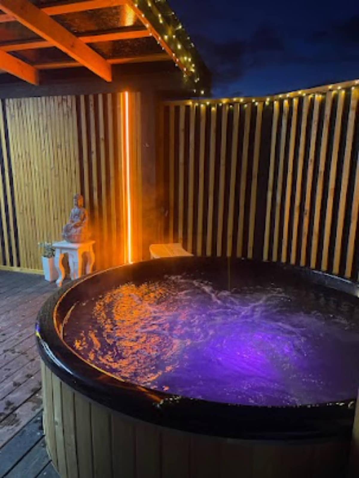 A wooden hot tub is illuminated by soft purple lights, creating a calming ambiance. The surrounding deck features wooden slats and draped fairy lights, enhancing the tranquil outdoor setting. A decorative statue is placed nearby, adding a serene touch to the relaxation area.