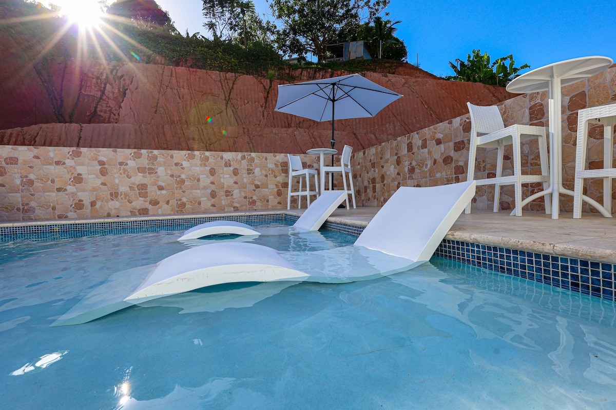 A tranquil swimming pool is featured with white lounge chairs partially submerged. A shaded table with two chairs is positioned nearby under a sun umbrella, set against a backdrop of lush greenery and a clear blue sky.