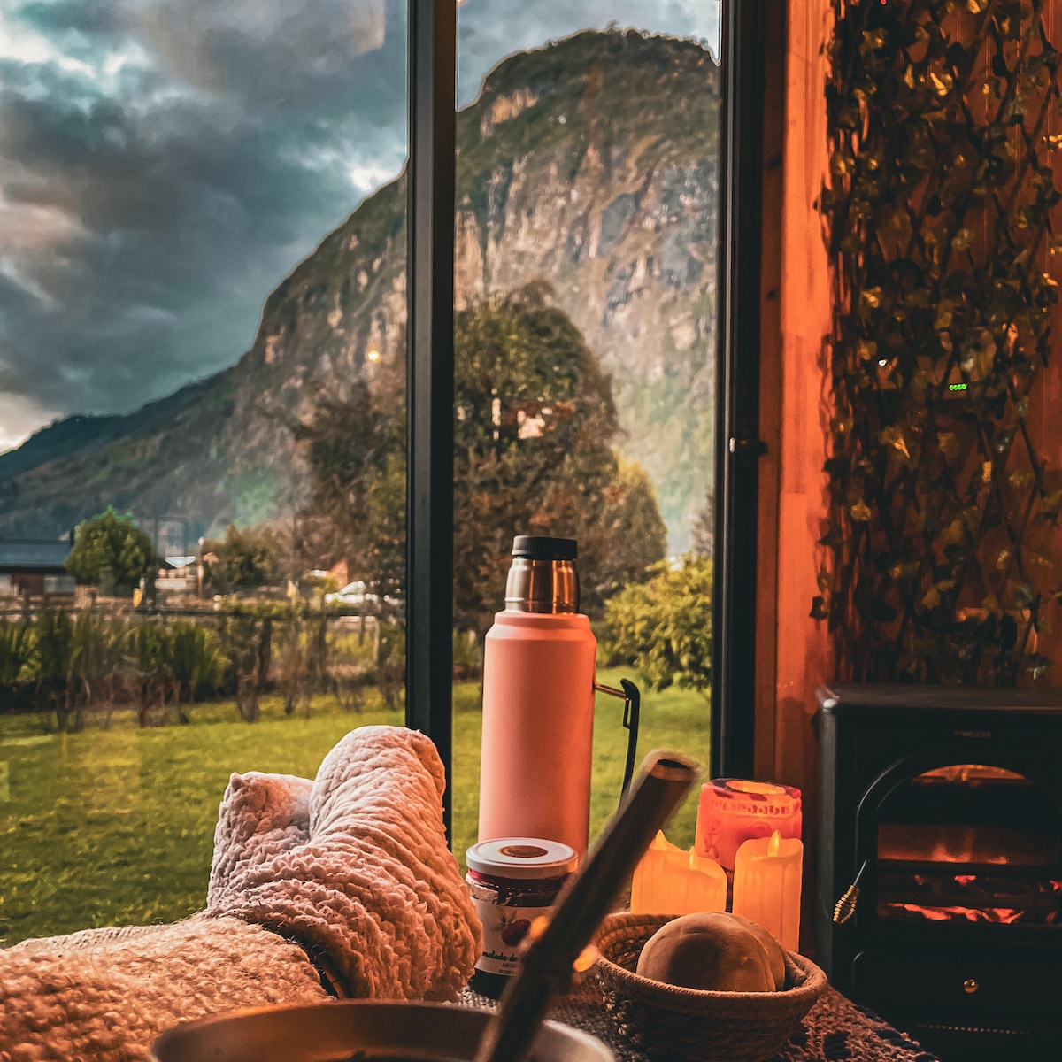 The image shows a cozy interior setting with a view of a mountainous landscape through large glass windows. A pair of feet in warm socks are positioned in the foreground, accompanied by a thermos, a steaming bowl, and softly glowing candles.