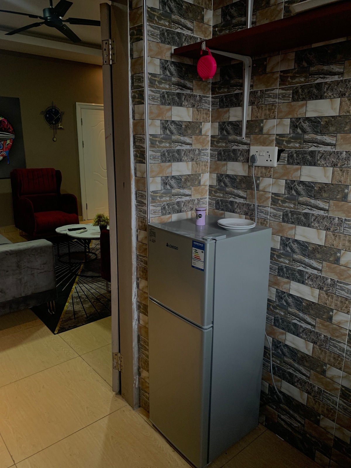 A compact refrigerator stands against a textured wall featuring a mix of light and dark tiles. A small dish is placed on top of the refrigerator. Nearby, a glimpse of a living area with a chair and coffee table can be seen.