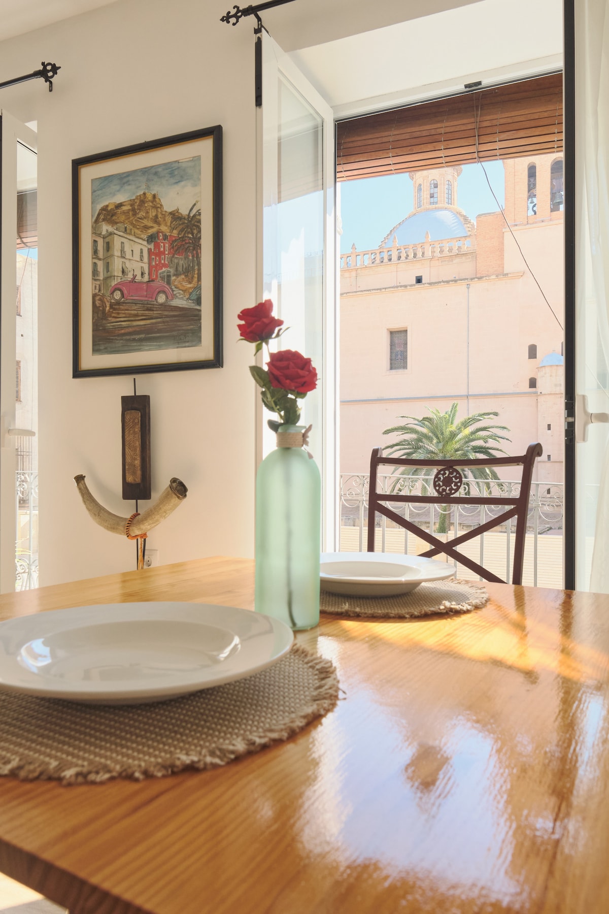 A dining area showcases a wooden table set with two plates and a slender glass vase containing roses. Light enters through large windows, framing a view of the historic cathedral and surrounding palm trees. A chair is positioned nearby, enhancing the inviting ambiance.