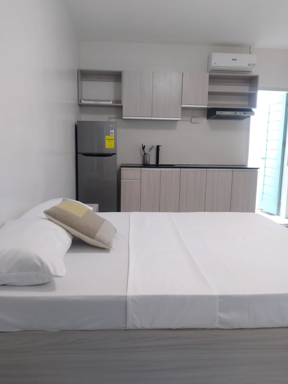 A spacious room is equipped with a large bed, a kitchen area featuring cabinets and a refrigerator. Natural light fills the space through a door leading to an outdoor area, which is partially visible. Neutral-colored furnishings create a calm environment.