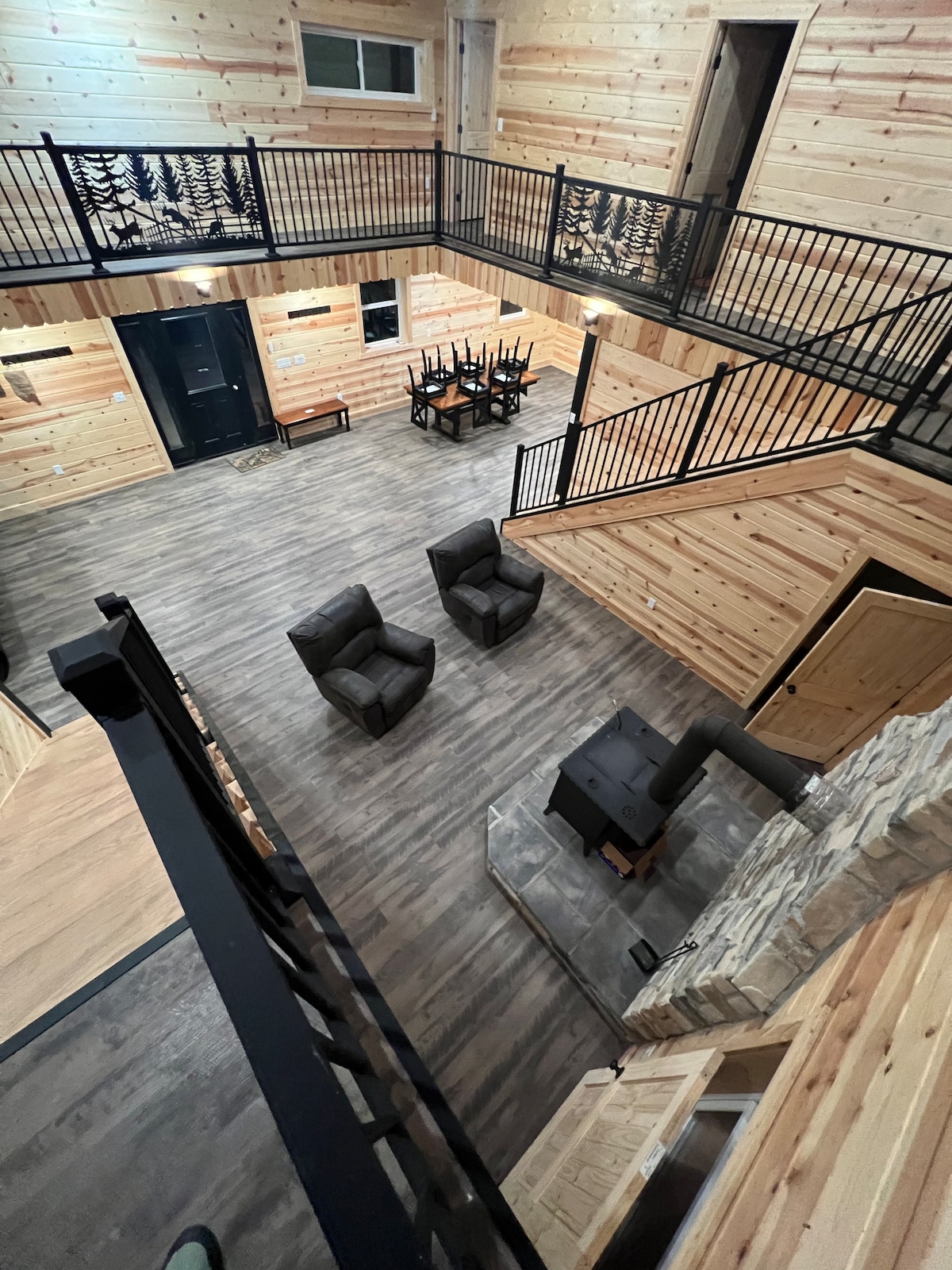 The interior showcases high wooden ceilings and walls, creating a warm cabin atmosphere. A spacious seating area features two armchairs positioned beside a wood-burning stove on a stone hearth. Dining chairs are visible in the background, framed by an open staircase.