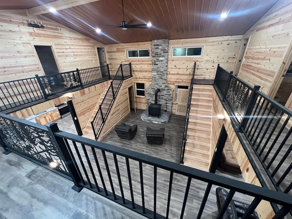 A spacious interior features an open layout with wooden walls and floors, showcasing two levels connected by elegant black railings. A central fireplace is surrounded by comfortable seating, with large windows allowing natural light to fill the area.